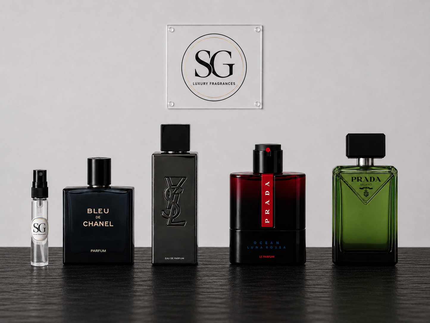 Five luxury perfume bottles arranged in a row on a dark tabletop against a light gray wall, with an SG Luxury Parfums sign centered above them. The bottles include a small clear sample spray, a black Chanel Bleu de Chanel, a black YSL logo bottle, a red-to-black Prada Eau de Parfum bottle, and a green Prada bottle.