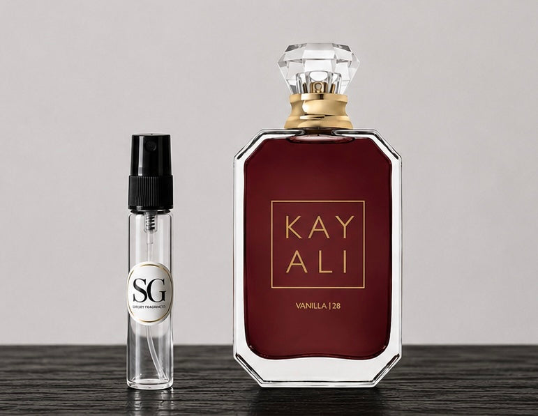 A large rectangular perfume bottle with a faceted clear cap and gold lettering reading 'KAY ALI' and 'VANILLA | 28' stands on a dark tabletop against a light gray background. Beside it is a small clear glass spray vial with a black spray top and a circular label reading 'SG'.
