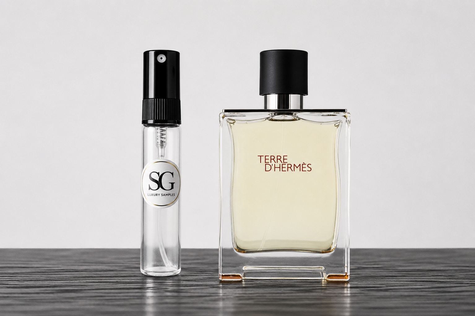 A clear glass perfume bottle with a black cap labeled 'TERRE D'HERMES' stands next to a smaller clear glass sample spray vial with a black spray top and a circular 'SG' label, set on a dark wooden surface against a light gray background.