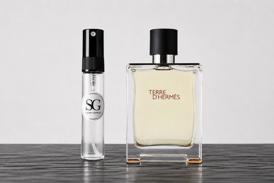 A clear glass perfume bottle with a black cap labeled 'TERRE D'HERMES' stands next to a smaller clear glass sample spray vial with a black spray top and a circular 'SG' label, set on a dark wooden surface against a light gray background.