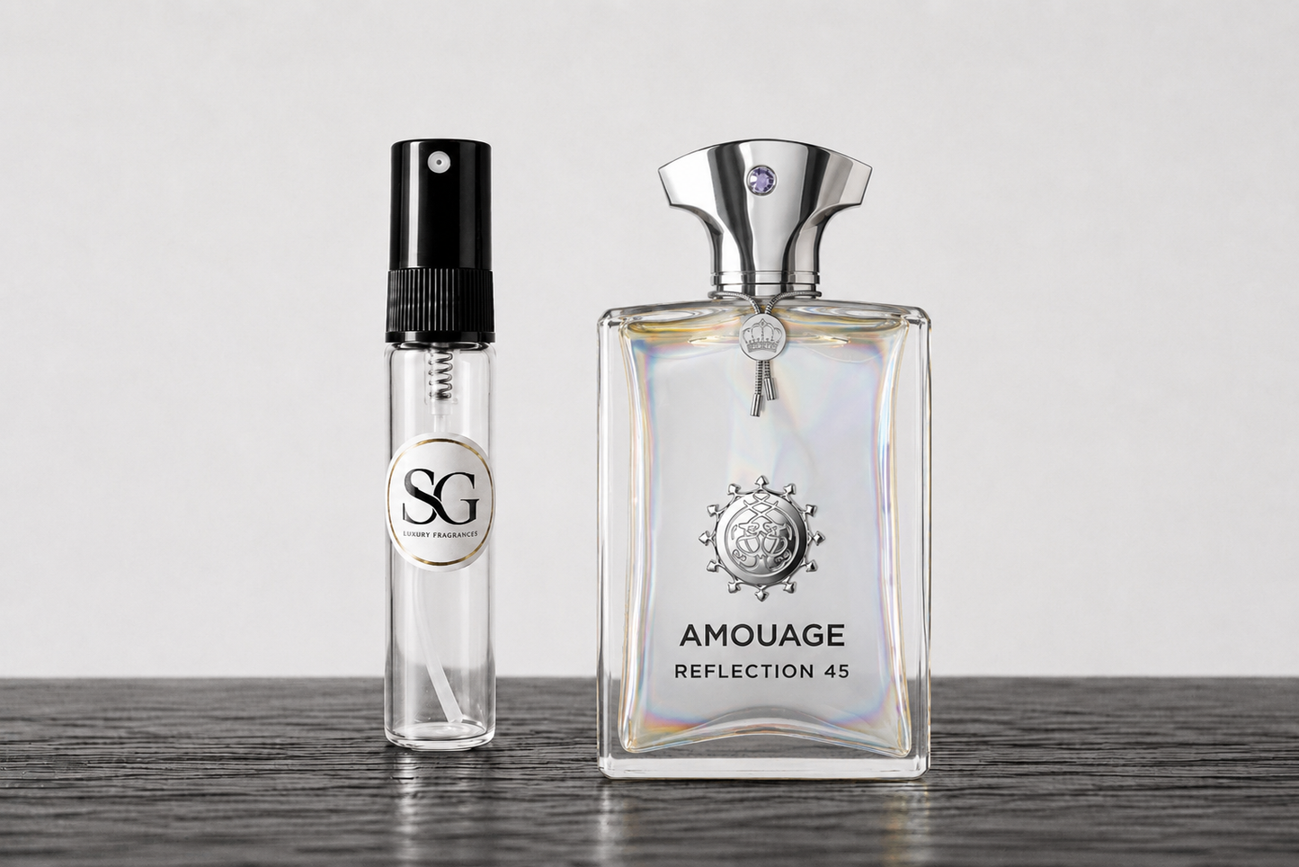 Amouage Reflection 45 perfume bottle next to a small clear glass sample spray vial with a black atomizer and a circular SG label, set on a dark wooden surface against a light gray background.