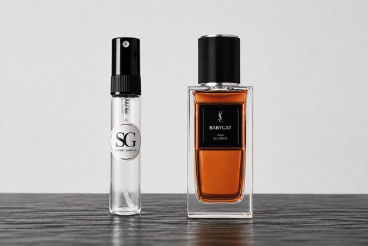 A clear glass perfume atomizer spray bottle with a black spray top and a circular silver 'SG' label, standing next to a larger rectangular clear glass bottle filled with amber liquid and labeled 'BABYCAT' by YSL, on a dark wood surface against a light gray background.