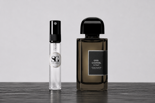 A small clear glass perfume sample spray bottle with a black spray top stands next to a larger dark brown glass perfume bottle labeled 'Oud Charvel Elixir' on a dark wooden surface against a light gray background.