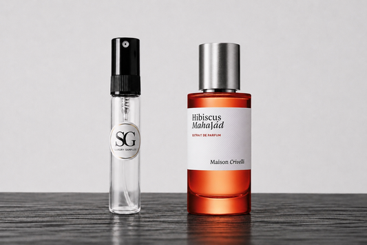 A clear glass perfume sample vial with a black spray top stands beside a larger amber glass bottle with a silver cap and a white label reading 'Hibiscus MahaJad' and 'Maison Orvelli'.