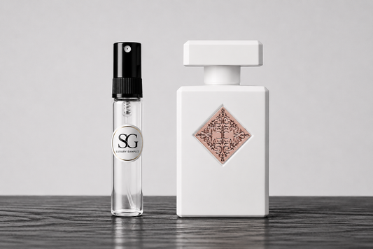 A clear glass perfume atomizer spray bottle with a black spray top and a circular silver label reading 'SG' stands next to a matte white rectangular perfume bottle with a decorative rose-gold diamond-shaped label. Both bottles are set on a dark wood surface against a light gray background.