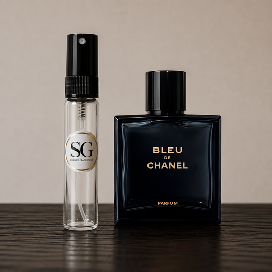 A clear glass perfume atomizer bottle with a black spray top and a circular gold-and-white 'SG' label stands beside a dark navy Chanel Bleu de Chanel perfume bottle on a wooden surface against a warm beige background.