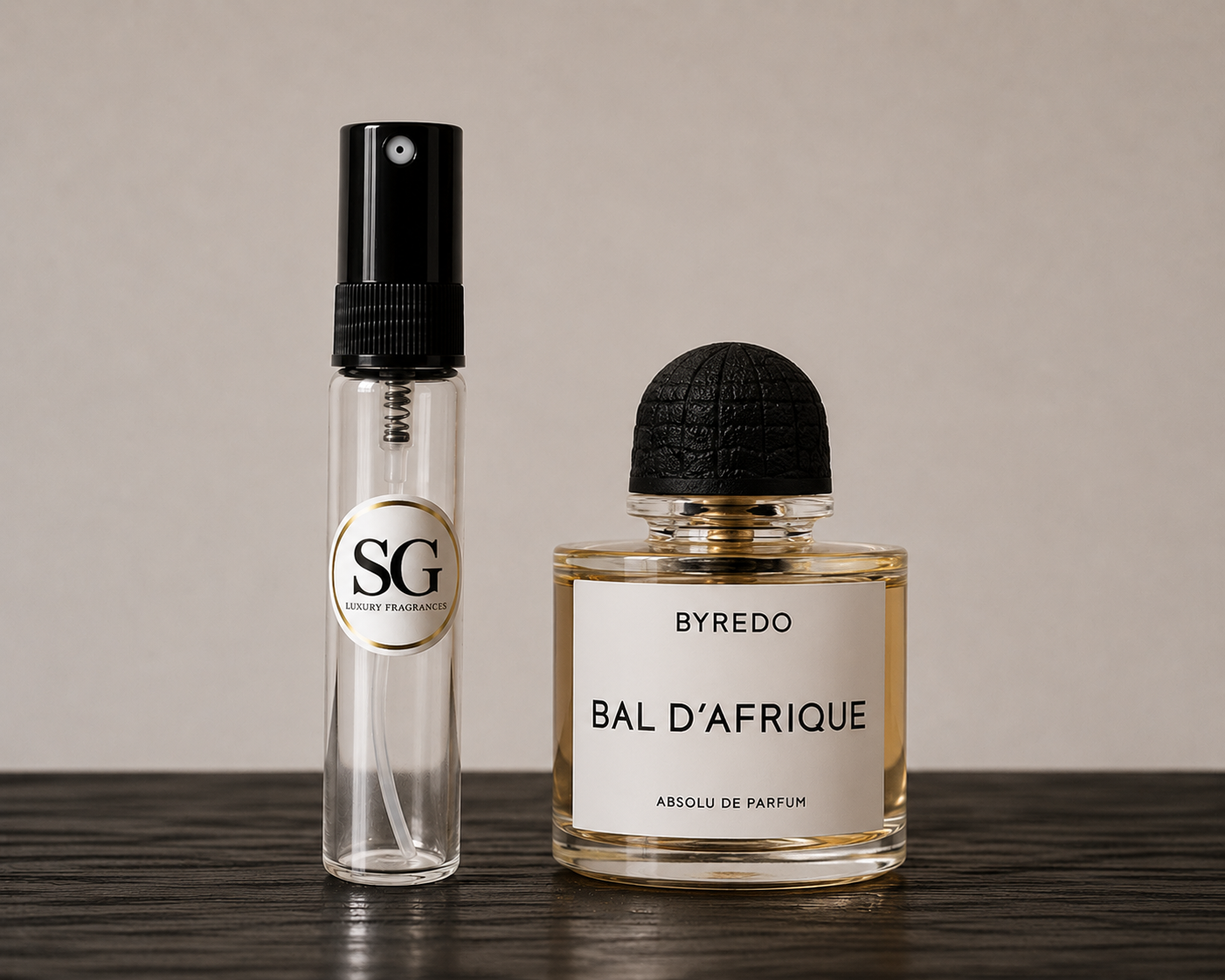 A clear cylindrical perfume atomizer spray bottle with a black spray top and a circular gold-and-black SG logo, standing beside a Byredo Bal D'Afrique perfume bottle with a black cap and white label on a dark wooden surface against a warm beige background.