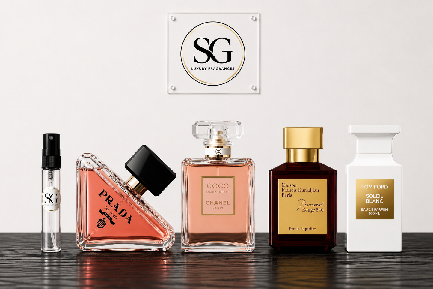 A lineup of five luxury perfume bottles on a dark wooden surface against a light gray wall, featuring a small clear spray vial, a pink Prada perfume bottle, a pink Chanel Coco perfume bottle, a dark brown Guerlain Affinity perfume bottle with gold detailing, and a white Tom Ford Solfi di Aglio bottle with a gold label. A square 'SG Luxury Fragrances' logo plaque is mounted above them.